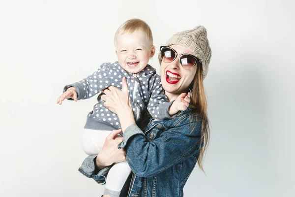 데님 재킷, 안경, 모자를 쓴 어린 소녀가 하얀 배경에 어린 아이를 안고 있다. 아이들과 개념 게임, 행복한 가족, 기쁨과 재미, 젊은 가족. — 스톡 사진