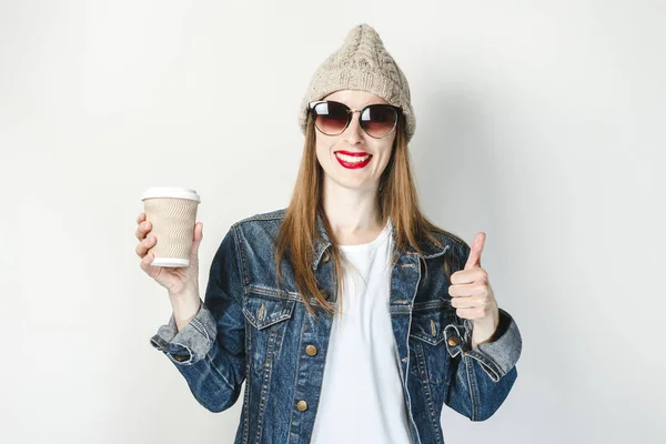 Una joven con una chaqueta de mezclilla, vasos y un sombrero sostiene una taza de café o té sobre un fondo blanco. Concepto desayuno, café en el trabajo, cafetería, recarga . — Foto de Stock