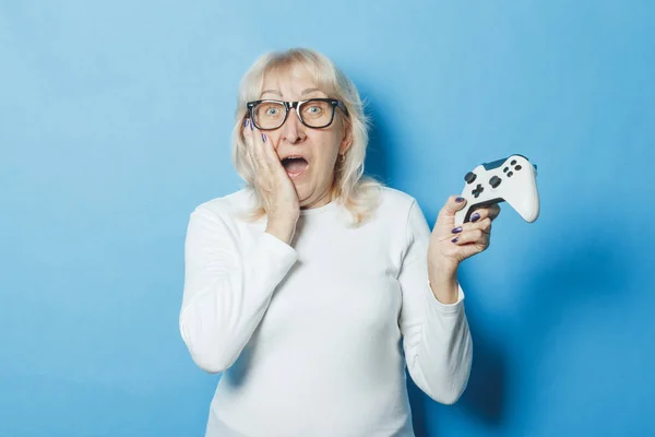 An old woman with a surprised face is holding a gamepad on a blue background. The concept of an old lady playing the console, game and entertainment for all.