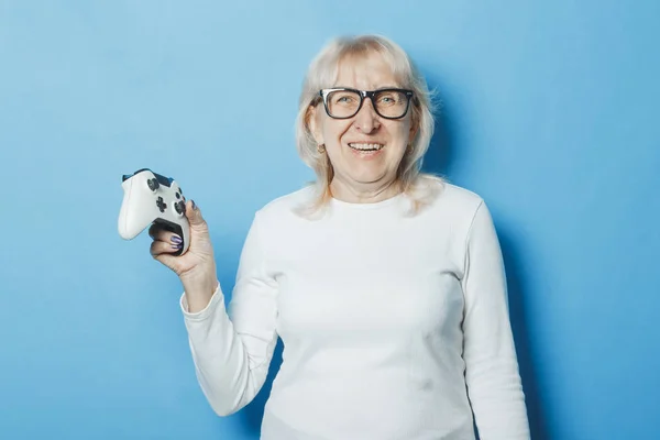 An old woman with a happy face is holding a gamepad on a blue background. The concept of an old lady playing the console, game and entertainment for all.