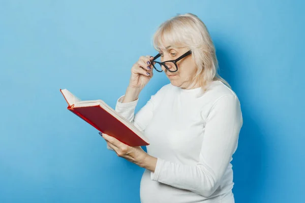 An old woman with glasses is reading a book on a blue background. Concept old lady reads books, education, book club.