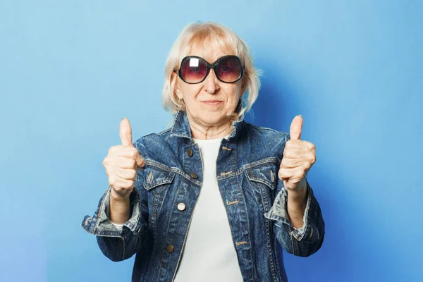 An old lady in a denim jacket and sunglasses is showing a gesture with her hands. Thumb up on a blue background. Concept fashionable grandma, old woman, funky action.