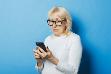 Gözlüklü yaşlı bir kadın elinde mavi bir arka plan üzerinde bir telefon tutar. Konsept Sms bildirimleri, yeni teknolojiler