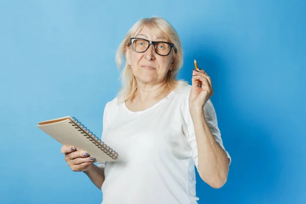 Velha senhora com óculos segura um caderno e caneta em suas mãos contra um fundo azul. Notas de conceito, palavras cruzadas, sudoku, lista de empilhamento, memória ruim — Fotografia de Stock