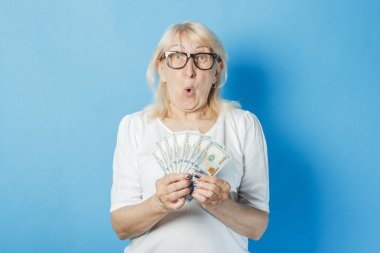 Old lady with glasses holds money in her hands against a blue background. Concept of wealth, winnings, savings, lottery, loan clipart