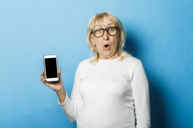 Gözlük takan şaşkın bir yüzü olan yaşlı kadın mavi bir arka plan üzerinde bir telefon tutuyor. Konsept Sms bildirimleri, yeni teknolojiler, promosyon ve satış, bildirim