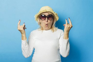 Hasır şapkalı ve güneş gözlüklü yaşlı kadın elleriyle mavi bir arka plana karşı bir jest yapıyor. Yaz başlangıcı kavramı, yaz saati, tatil, kamp