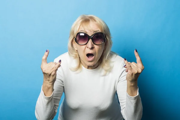 Old woman wearing glasses with a surprised face makes a gesture with her hands against a blue background. Concept rock, old rocker, goat gesture