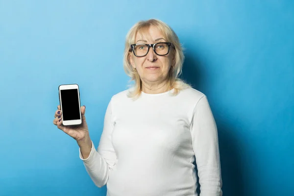 Old woman with glasses holds a telephone in hand on a blue background. Concept SMS notifications, new technologies