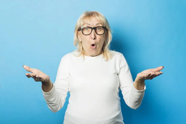 Old woman wearing glasses with a surprised face makes a gesture with her hands against a blue background. Concept of surprise, shock, unexpected news
