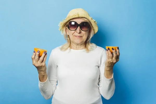 Старуха в соломенной шляпе и солнечных очках держит апельсин на синем фоне. Наступление лета, летнее время, отпуск, кемпинг — стоковое фото
