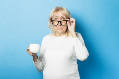 Mavi arka planda bir bardak kahve tutan gözlüklü yaşlı bir kadın. Konsept kahve dükkanı, şarj, kahve enerjisi