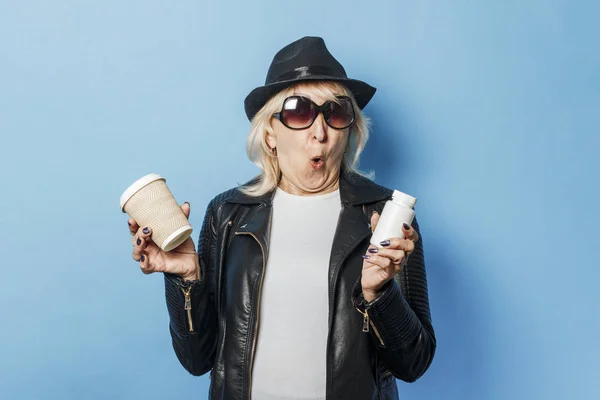 Old lady wearing glasses, a leather jacket and a hat is holding a glass with coffee and a bottle with pills on a blue background. Concept of abuse of relaxants, choice between health and harm