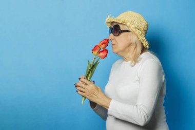 Hasır şapkalı ve güneş gözlüklü yaşlı kadın mavi bir arka plan üzerinde bir buket lale tutuyor. İlkbahar, yaz saati, tatil, kamp başlangıcı kavramı