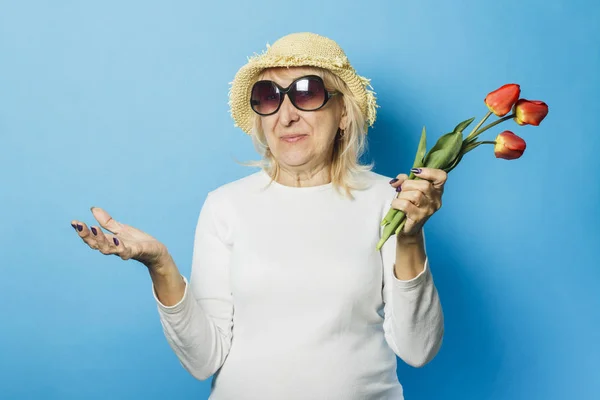 Старуха в соломенной шляпе и солнечных очках держит букет тюльпанов на синем фоне. Начало весны, лето, отпуск, кемпинг — стоковое фото