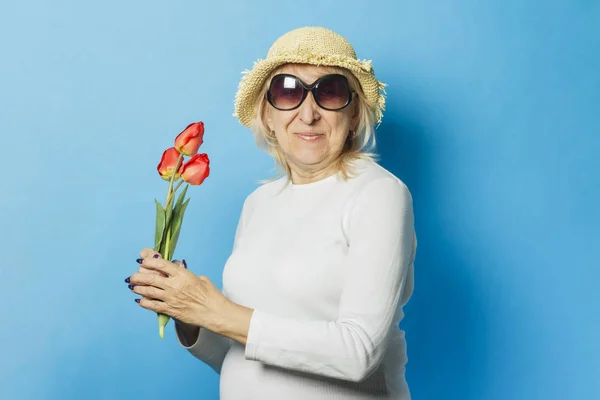 Старуха в соломенной шляпе и солнечных очках держит букет тюльпанов на синем фоне. Начало весны, лето, отпуск, кемпинг — стоковое фото