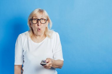 Şaşırmış suratlı yaşlı bir kadın mavi arka planda bir televizyon kumandası tutuyor. Yaşlılar için konsept televizyon, yeni teknolojilerin çalışması, Tv dizileri, kablolu televizyon, yüksek kalite
