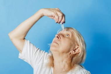 Yaşlı bir kadın mavi bir arka plan karşı gözlerine bir damla damla damlayor. Göz tedavisi kavramı, görme nin iyileştirilmesi, görme nin zayıflığı