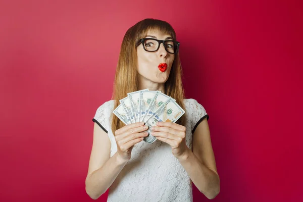 Junge Frau mit überraschtem Gesicht hält Geld in den Händen vor dunkelrotem Hintergrund. Konzept reich, Gewinne, Lotterie, Kredit, Bestätigung — Stockfoto