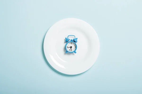 Reloj despertador azul se encuentra en una placa blanca sobre un fondo azul. Concepto de dieta, horario de comidas, pérdida de peso. Piso tendido, vista superior — Foto de Stock