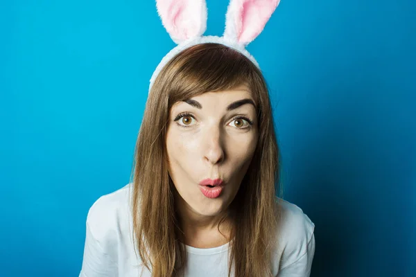 Mujer joven en orejas de conejo con una cara sorprendida sobre un fondo azul. Concepto Pascua — Foto de Stock