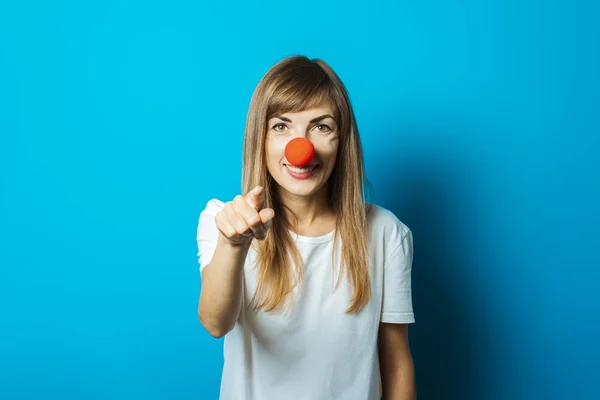 Piękna młoda kobieta w białym T-shirt i czerwony nos clown uśmiecha się i wskazuje palcem na niebieskim tle. Impreza koncepcyjna, kostium, czerwony nos dzień — Zdjęcie stockowe