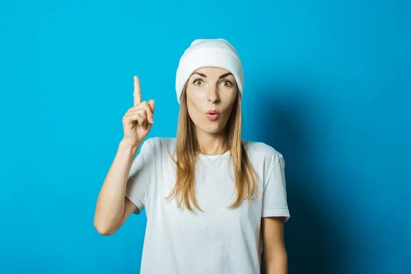 Mujer joven con un sombrero blanco y una camiseta con una cara sorprendida sobre un fondo azul — Foto de Stock