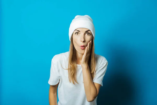 Mujer joven con un sombrero blanco y una camiseta con una cara sorprendida sobre un fondo azul — Foto de Stock