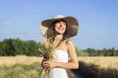 Beyaz elbiseli ve şapkalı genç ve güzel bir kadın sütlü tarlaların arasındaki yolda kır çiçekleriyle dolu bir buket tutuyor. Açık hava eğlencesi kavramı, köy gezisi.