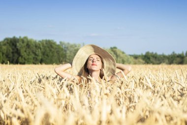 Geniş kenarlı şapkalı genç kadın buğday tarlasını dikizliyor.