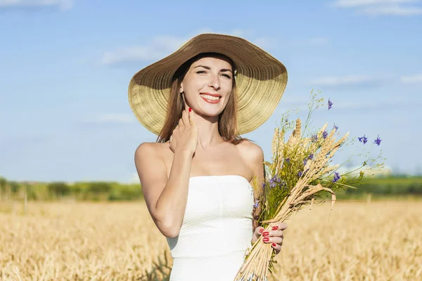 Joven mujer hermosa en un vestido blanco y un sombrero sostiene un ramo de flores silvestres en un campo de mijo. Concepto de recreación al aire libre, un viaje al pueblo — Foto de Stock