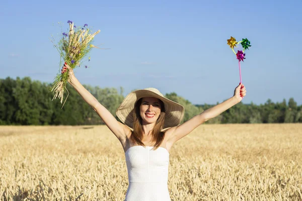Mulher bonita nova em um vestido branco e chapéu mantém um buquê com flores silvestres e uma viseira de brinquedo no campo de um milf. Conceito de recreação ao ar livre, uma viagem para a aldeia — Fotografia de Stock