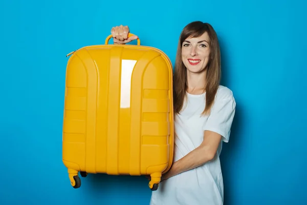 Die schöne junge Frau hält einen gelben Plastikkoffer vor blauem Hintergrund. Konzept Reisegepäck, Reise, Urlaub — Stockfoto