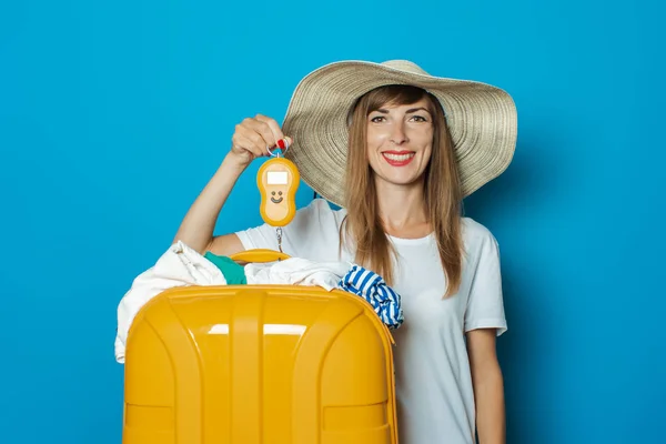 Die schöne junge Frau mit Hut wiegt einen überfüllten gelben Plastikkoffer auf blauem Grund. Konzeptgepäck, Übergepäck, Gepäckgewicht — Stockfoto