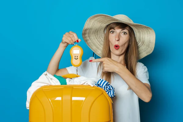 Die schöne junge Frau mit dem überraschten Gesicht im Hut wiegt einen überfüllten gelben Plastikkoffer auf blauem Grund. Konzeptgepäck, Übergepäck, Gepäckgewicht — Stockfoto