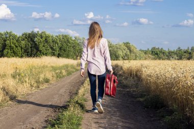 Bir bavul ile güzel genç kadın arka planda buğday tarlaları arasında bir yol boyunca yürüyor. Seyahat konsepti, tatil