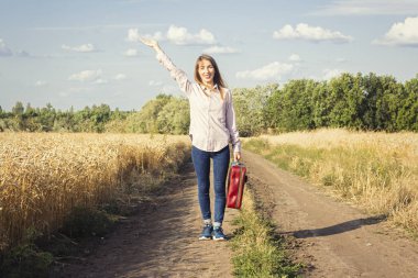 Beautiful young woman with a suitcase on the road between wheat fields in the background. Travel concept, vacation