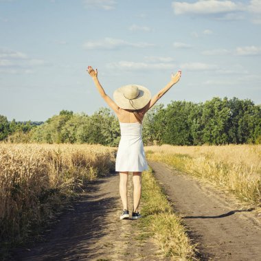 Beyaz elbiseli ve şapkalı güzel genç bir kadın buğdaylı iki tarla arasında yol boyunca yürüyor. Açık hava eğlencesi, köy gezisi, tatil.