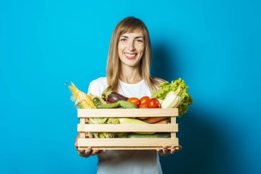 Genç bir kadın mavi arka planda taze sebzelerle dolu bir kutu tutuyor. İyi hasat kavramı, doğal ürün