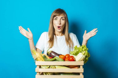 Şaşırmış yüzlü genç bir kadın mavi arka planda taze sebzelerle dolu bir kutu tutuyor. İyi hasat kavramı, doğal ürün