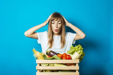 Şaşırmış yüzlü genç bir kadın mavi arka planda taze sebzelerle dolu bir kutu tutuyor. İyi hasat kavramı, doğal ürün