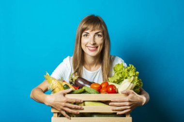 Genç bir kadın mavi arka planda taze sebzelerle dolu bir kutu tutuyor. İyi hasat kavramı, doğal ürün