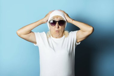 Şapkalı ve gözlüklü komik yaşlı kadın mavi arka planda el hareketi yapıyor. Modern tarzda havalı bir büyükanne kavramı.