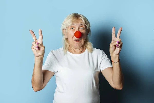 Old woman with a red clown nose makes hand gestures on a blue background. Red nose day concept, clown, party
