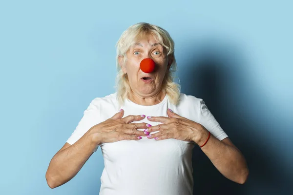 Old woman with a red clown nose makes hand gestures on a blue background. Red nose day concept, clown, party