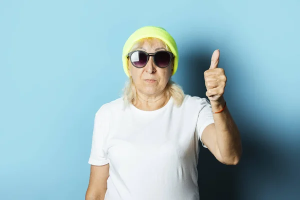Забавная старушка в шляпе и очках, делает жест рукой на синем фоне. Концепция прохладно стильная бабушка, современный стиль — стоковое фото