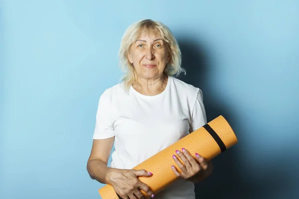 Old woman holds a yoga mat on a blue background. Yoga concept for old people, gymnastics and Pilates