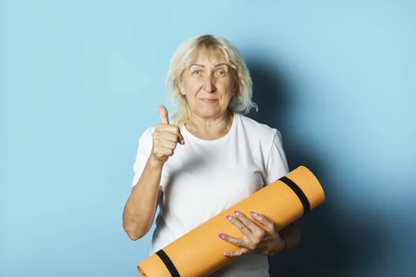 Old woman holds a yoga mat on a blue background. Yoga concept for old people, gymnastics and Pilates