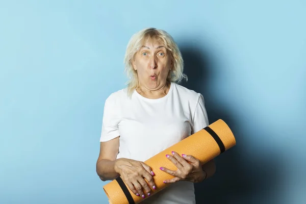 Old woman holds a yoga mat on a blue background. Yoga concept for old people, gymnastics and Pilates