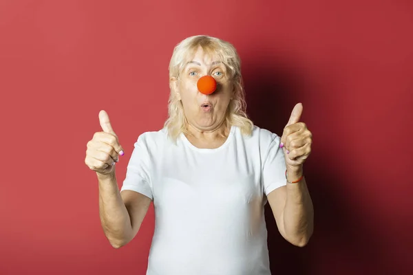 Funny old woman with a red nose of a clown on a pink background. Concept red nose day, holiday, party, clown